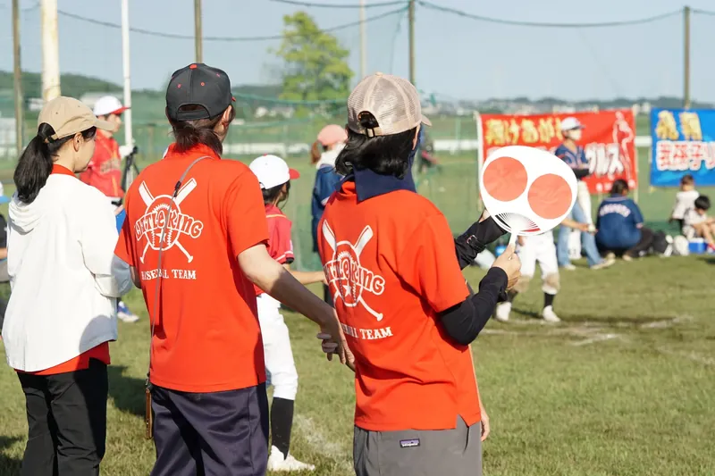 チームのTシャツを着てグラウンドで子どもたちを見守る保護者たち